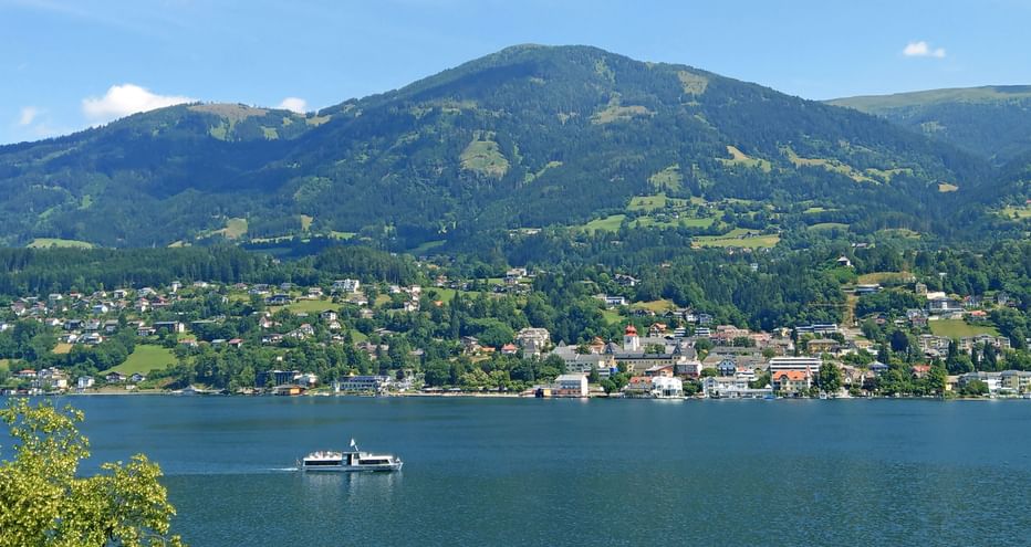 Passenger ferry crossing a Carinthian lake with a town on the shore and green mountains in the background under blue sky.