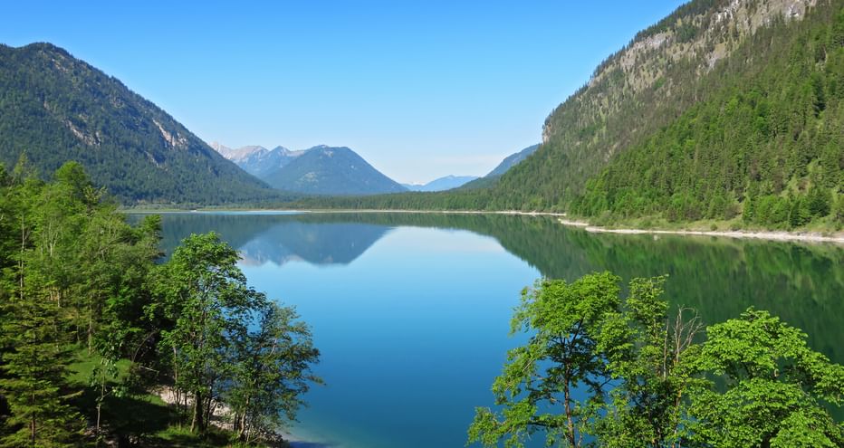 Sylvensteinstausee umgeben von bewaldeten Bergen mit ruhigem blauen Wasser, das Himmel und Gipfel in den bayerischen Alpen spiegelt.