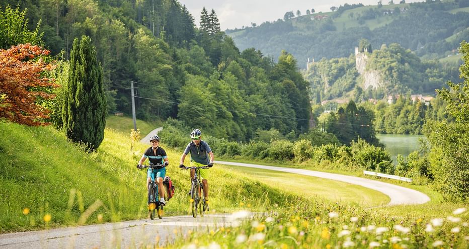 Zwei Radfahrer auf asphaltiertem Radweg, umgeben von Wiesen, im Hintergrund Burg Losenstein und bewaldete Hügel