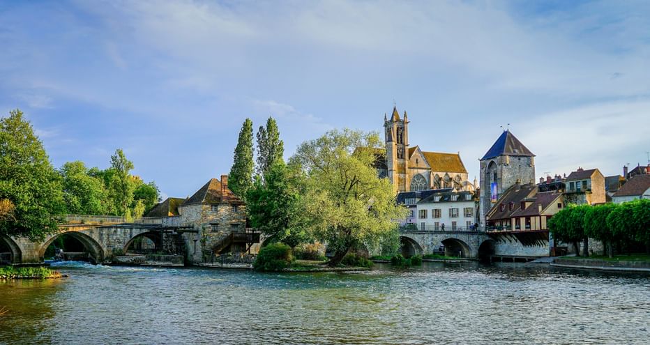 View of Moret-sur-Loing with stone bridge crossing the Loing river, historic church with tower, and traditional buildings under blue sky.