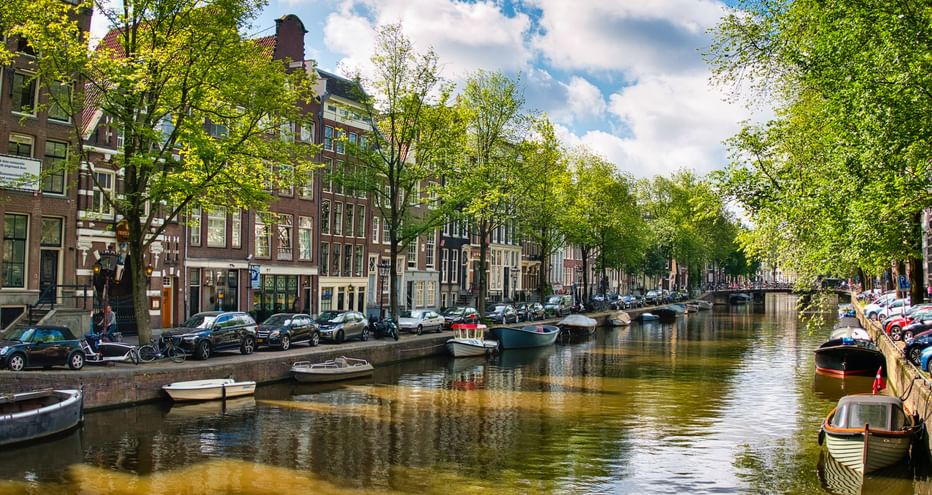 Amsterdamer Gracht gesäumt von traditionellen holländischen Häusern, parkenden Autos und Booten. Bäume rahmen beide Seiten unter blauem Himmel.