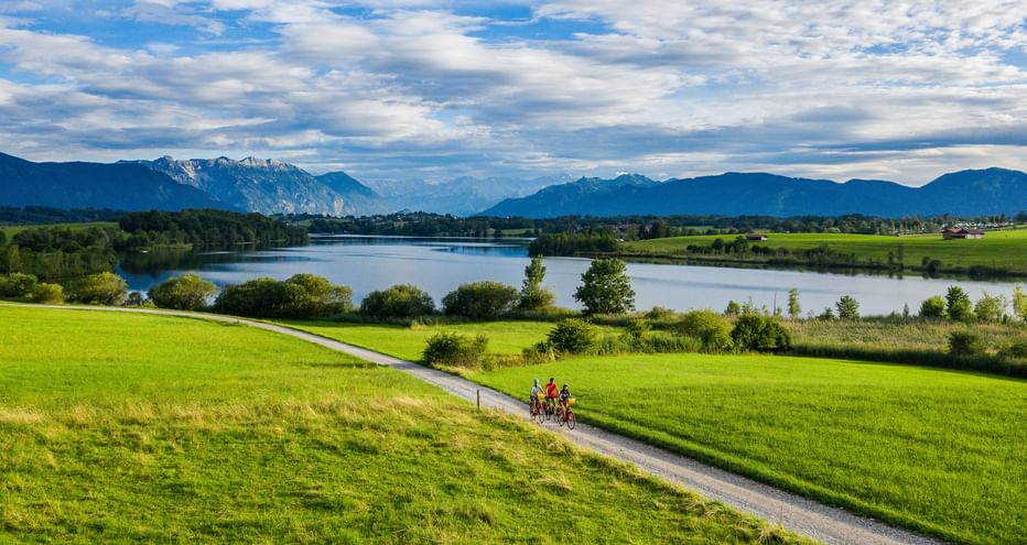 Two cyclists on a path through green meadows near Riegsee lake in Bavaria, with mountains and blue sky in the background.