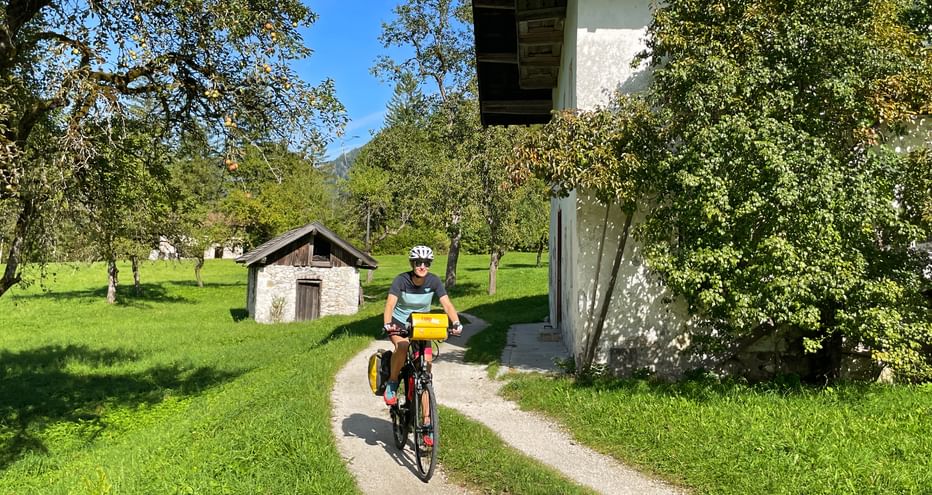 Cyclist with yellow panniers riding on a paved path through green meadows near Reit on the Tauern Radweg, with traditional buildings and trees.