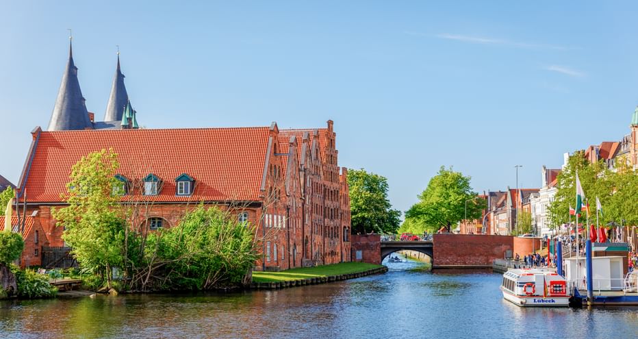 Rote Backstein-Salzspeicher am Kanal in Lübeck mit Kirchtürmen, Brücke und Ausflugsboot unter blauem Himmel.