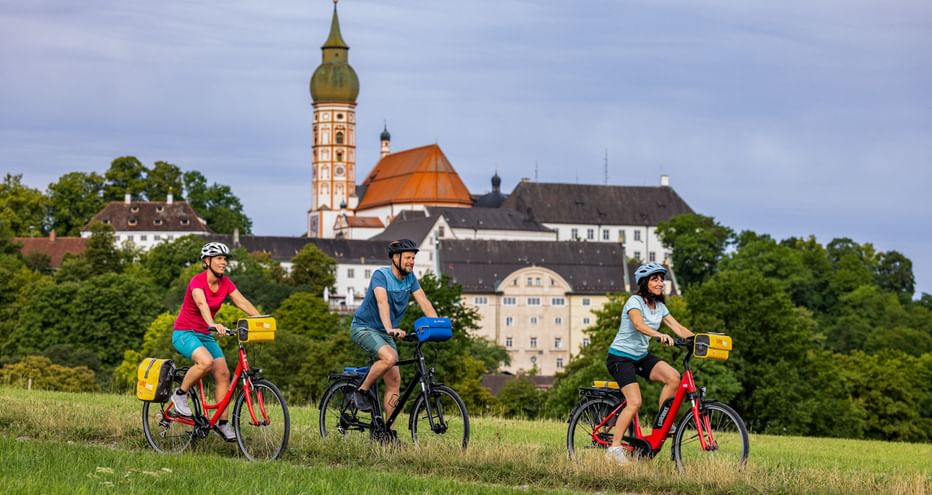 Three cyclists riding through a green meadow with Andechs Monastery visible on a hill in the background, featuring a baroque church tower.