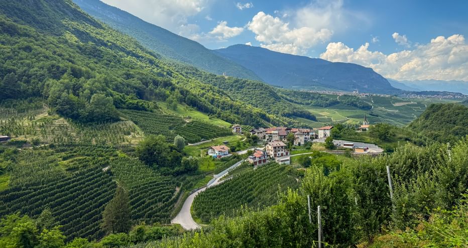 Kleines Dorf inmitten terrassierter Weinberge in einem Bergtal. Grüne Hänge mit Weinreben unter blauem Himmel mit weißen Wolken.