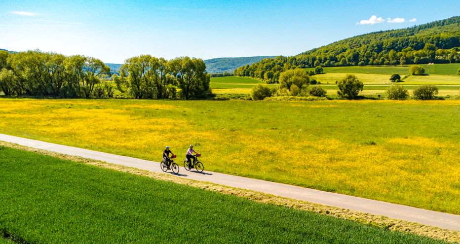 Two cyclists riding on a paved path through green and yellow meadows in the Weserbergland region with forested hills in the background.