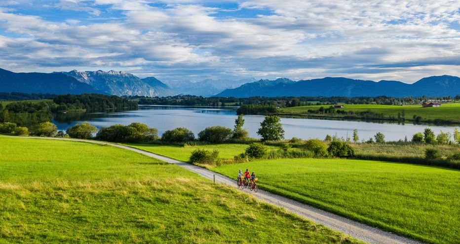 Two cyclists on a path through green meadows near Riegsee lake in Bavaria, with mountains in the background under a cloudy blue sky.