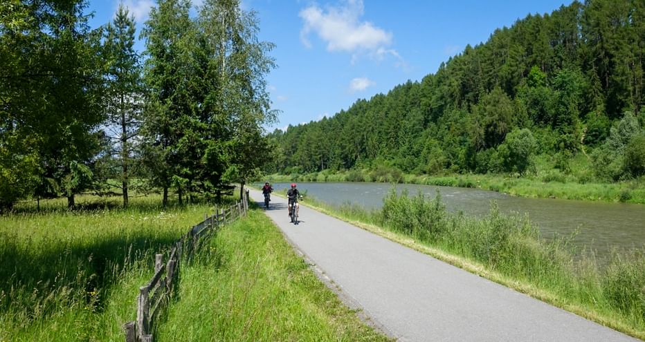 Cyclists on paved path along Dunajec River with wooden fence, green meadows, and forested hills under blue sky.