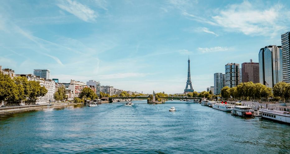 View along the Seine River in Paris with the Eiffel Tower in the distance, modern buildings on both banks, and boats on the water under a blue sky.