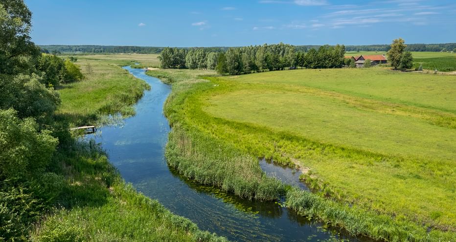 Luftaufnahme eines blauen Flusses, der sich durch grüne Wiesen bei Wojnowo-Kruttina in Masuren schlängelt, mit Bäumen und Bauernhaus.