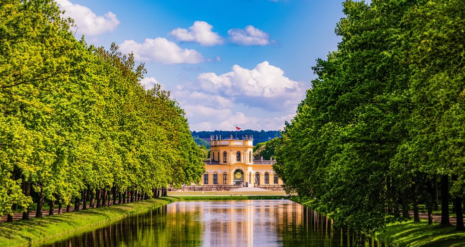 Gelbes barockes Orangerie-Schloss in Kassel, eingerahmt von baumgesäumtem Kanal mit Spiegelungen. Blauer Himmel mit weißen Wolken über der symmetrischen Szene.