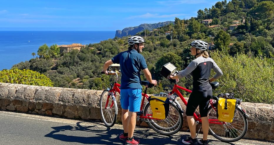 Two cyclists with helmets and red touring bikes with yellow panniers studying a map on a coastal road in Mallorca, overlooking the blue sea.