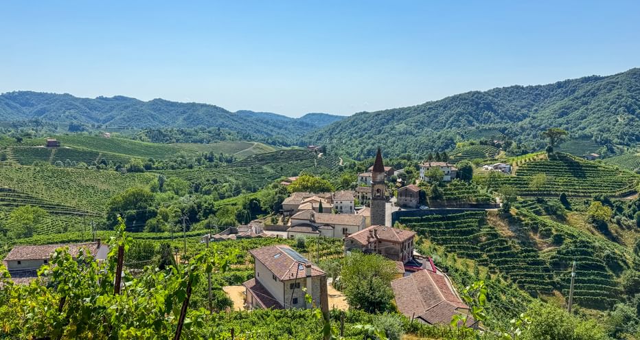 Kleines italienisches Dorf mit Kirchturm, umgeben von terrassierten Prosecco-Weinbergen bei Conegliano, mit sanften Hügeln im Hintergrund.