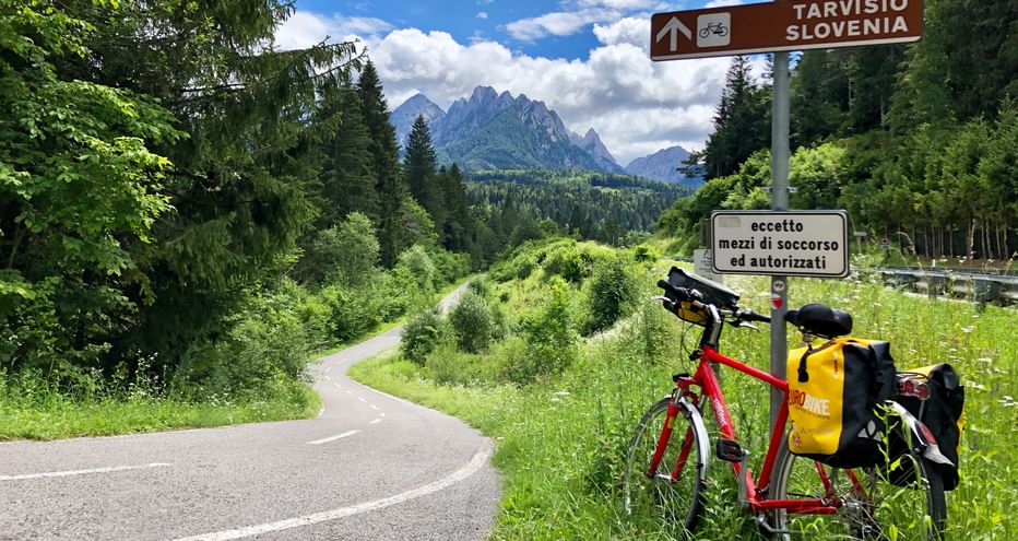 Cycle break with bike leaning against a road sign, with beautiful cycle path in the foreground and the Carnic Alps in the background