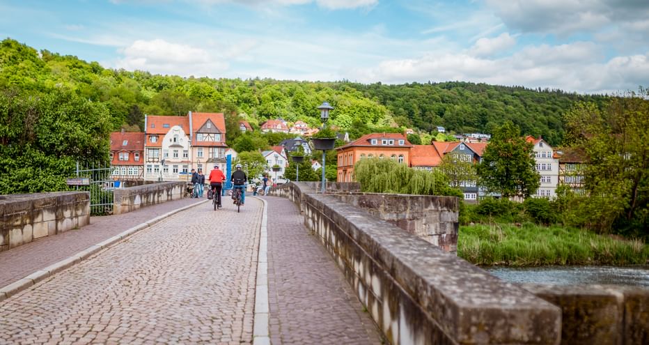 Radfahrer überqueren historische Steinbrücke in Hann. Münden mit bunten Fachwerkhäusern und grünen Hügeln im Hintergrund.