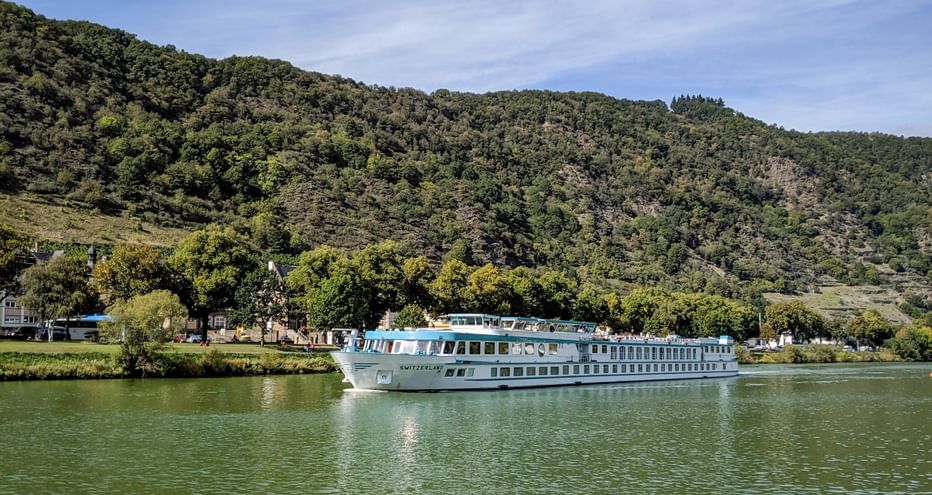 MS Switzerland Flusskreuzfahrtschiff fährt auf ruhigem grünen Wasser mit bewaldeten Hügeln im Hintergrund unter teilweise bewölktem Himmel.