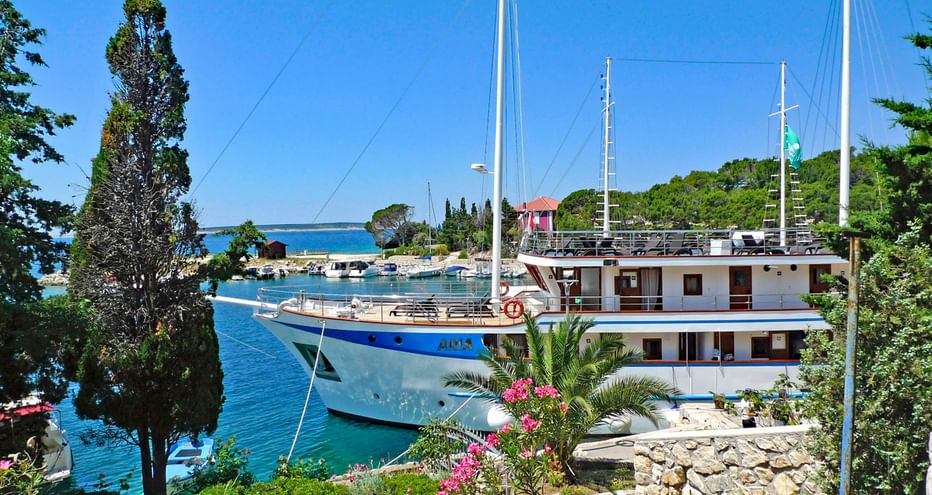 White two-deck MS Aria ship with blue hull and two masts moored in a Mediterranean harbor. Pink flowers and cypress trees frame the view.