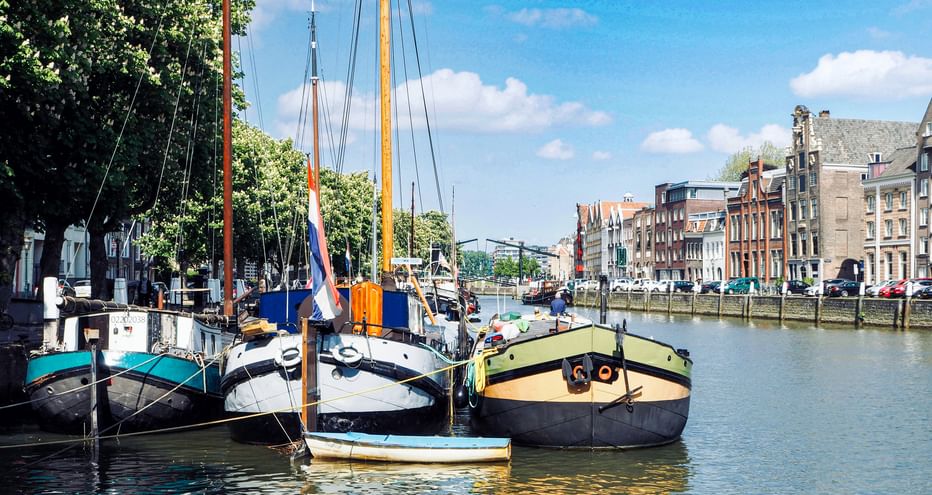 Traditionelle Segelboote im Kanal von Dordrecht mit historischen niederländischen Gebäuden am Ufer unter blauem Himmel.