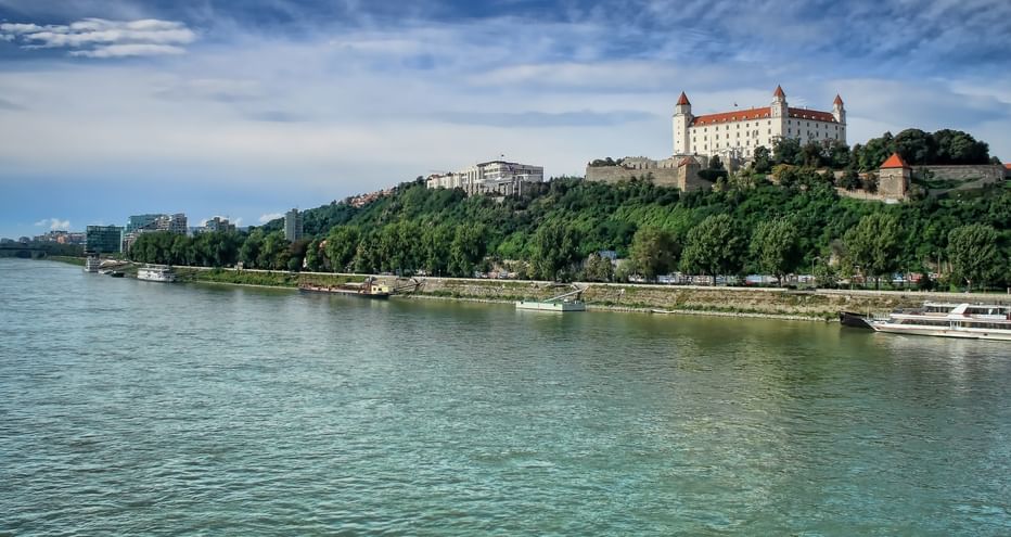Bratislava Castle with white walls and red roofs on a green hill above the Danube River. Ships are moored along the tree-lined riverbank.