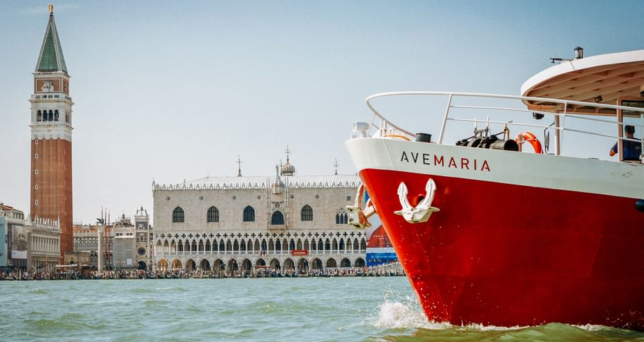 Rot-weißes Ave Maria Schiff in der Lagune von Venedig mit Markusturm und Dogenpalast im Hintergrund unter klarem blauen Himmel.