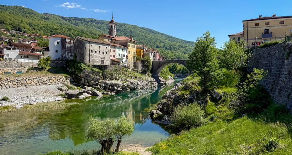 Historic stone bridge spanning a green river in Kanal ob Soči, Slovenia, with colorful buildings and forested hills in the background.