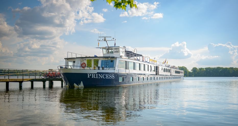 River cruise ship MS Princess moored at a wooden pier on calm water. The white vessel reflects in the water under a blue sky with clouds.