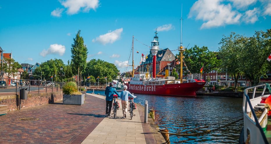 Zwei Radfahrer fahren entlang eines gepflasterten Uferwegs mit rotem Feuerschiff und Leuchtturm im Hafen unter blauem Himmel mit weißen Wolken.