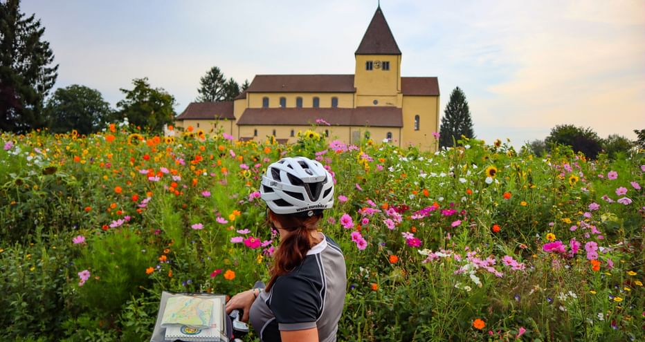 Eine Radfahrerin steht vor einer Blumenwiese und einer Kirche