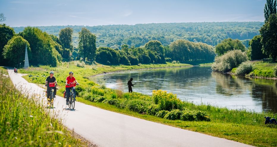 Two cyclists in red shirts riding along paved path beside Weser River. Green landscape with trees and hills in background under blue sky.