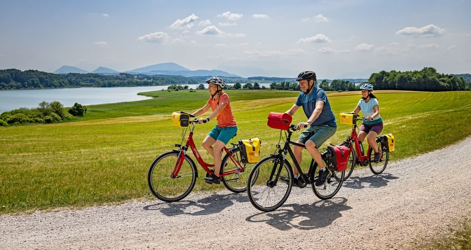 Drei Radfahrer mit Tourenrädern auf einem Schotterweg durch grüne Felder. Im Hintergrund sind ein See und Berge unter blauem Himmel sichtbar.