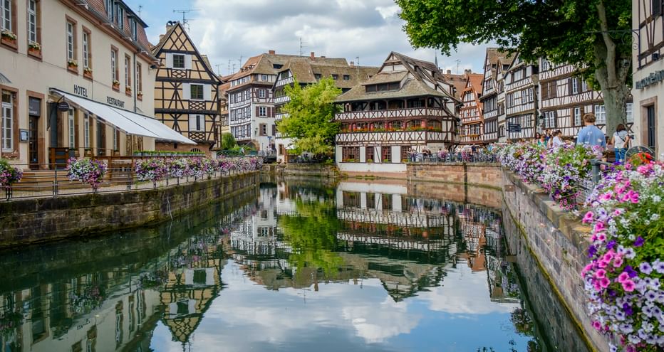 Kanal in La Petite France, Straßburg, gesäumt von rosa Blumen. Traditionelle Fachwerkhäuser spiegeln sich im ruhigen Wasser unter bewölktem Himmel.