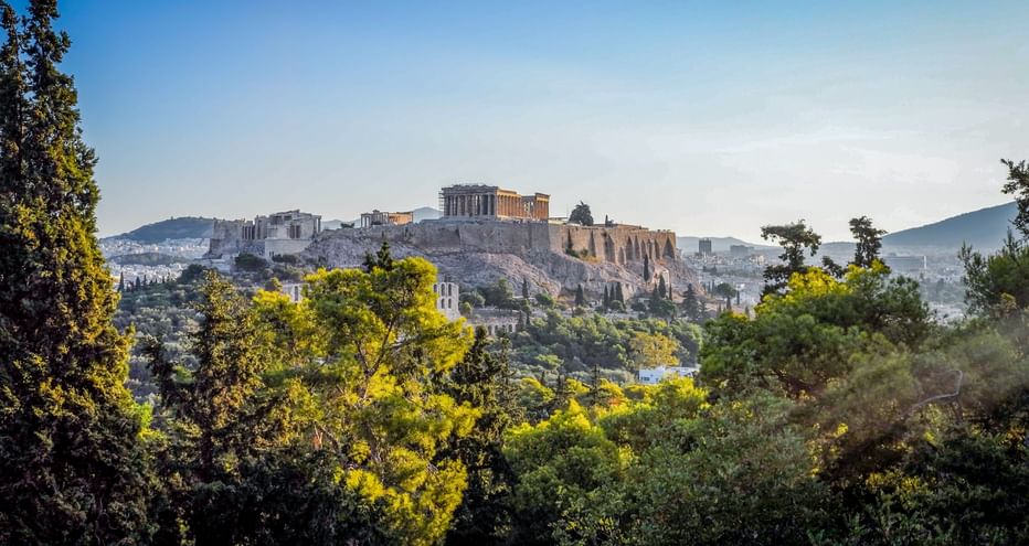 Blick auf die Akropolis mit dem Parthenon-Tempel in Athen, Griechenland. Antike Ruinen auf felsigem Hügel umgeben von grünen Bäumen unter klarem Himmel.