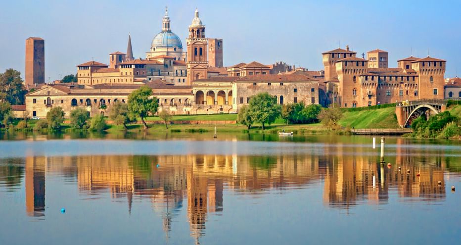 Historische Skyline von Mantua mit Domkuppel und Glockenturm spiegelt sich in ruhigem Wasser. Grüne Bäume säumen das Ufer mit terrakottafarbenen Gebäuden.