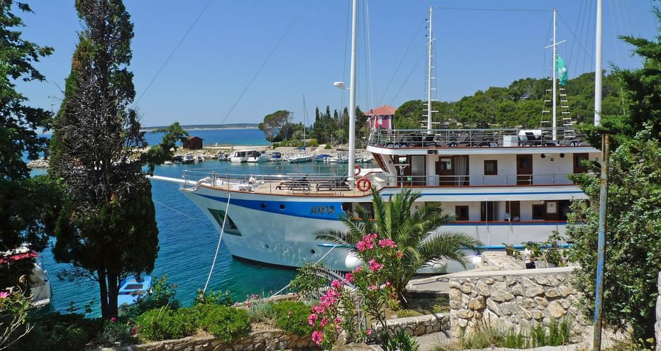 Weiße und blaue Motoryacht MS Aria mit zwei Masten im Mittelmeerhafen. Steinmauern mit rosa Blumen rahmen den Blick auf das türkisfarbene Wasser.