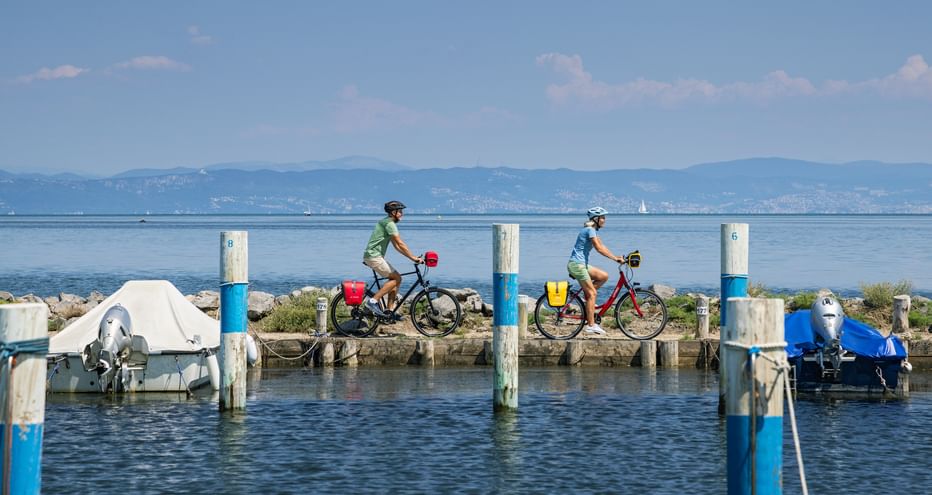 Two cyclists riding along a wooden pier in Grado harbor with boats moored on both sides, mountains visible in the background under blue sky.