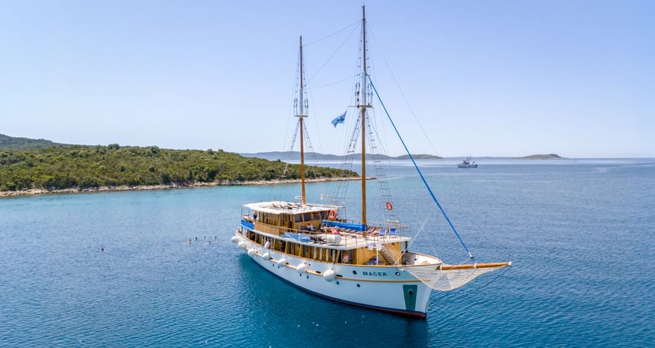 White two-masted sailing vessel MS Macek anchored in calm turquoise waters near a green forested coastline under clear blue sky.