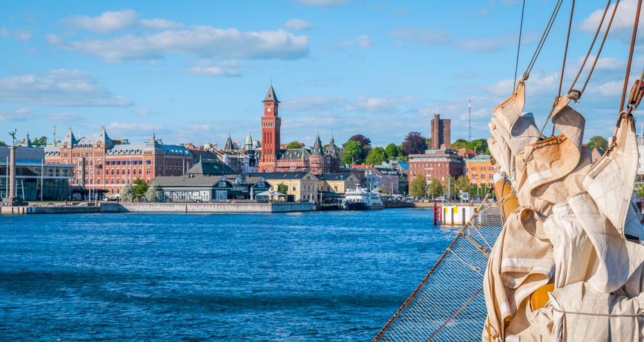 View of Helsingborg waterfront from a sailing ship. Red brick tower and historic buildings line the harbor under blue sky with white clouds.