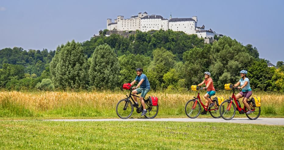 Three cyclists riding on a path through green meadows with Hohensalzburg Fortress on a wooded hill in the background under blue sky.