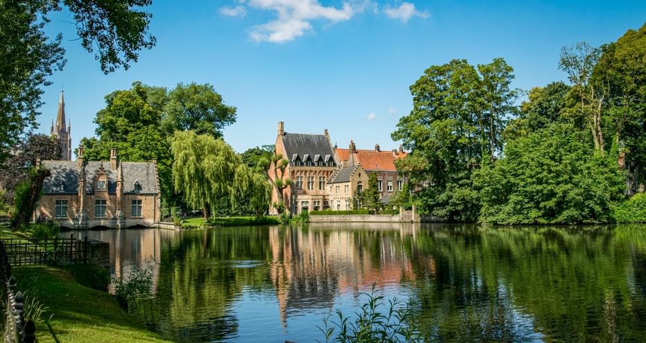 Minnewater Park in Bruges showing a tranquil lake reflecting historic buildings with red roofs and a church spire, surrounded by lush green trees.