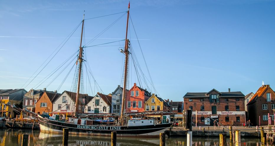 Traditionelles Zweimaster-Segelschiff im Hafen von Undine vor bunten historischen Gebäuden unter blauem Himmel.