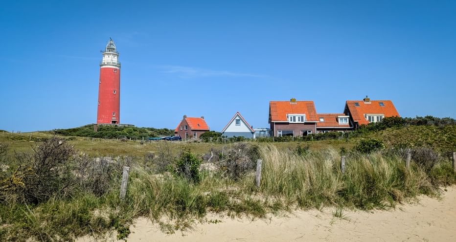 Red lighthouse on Texel island with traditional Dutch houses with orange roofs, surrounded by dune grass and sandy beach under blue sky.