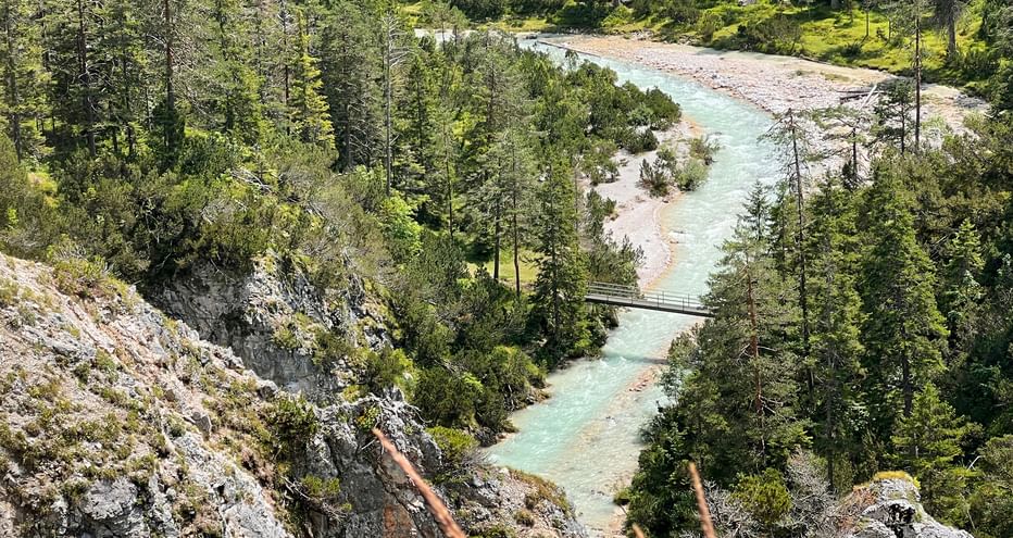Turquoise Isar river flowing through dense forest with rocky banks. A small footbridge crosses the river in the Munich lakes region.