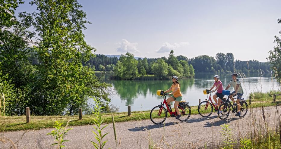 Drei Radfahrer fahren auf einem Schotterweg entlang eines ruhigen Sees in Kärnten. Grüne Bäume rahmen die Szene mit bewaldeten Hügeln im Hintergrund.