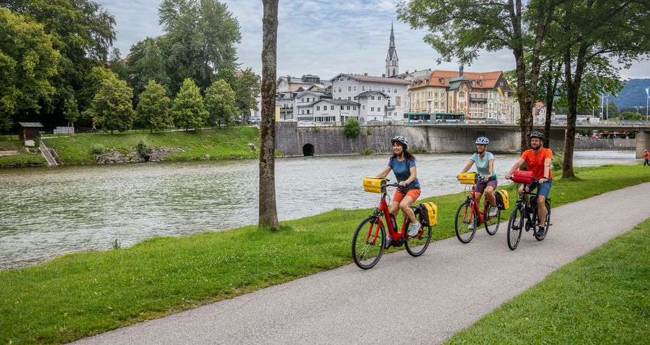 Three cyclists with yellow panniers riding along the Isar River in Bad Tölz. Historic buildings and a church spire visible across the river.