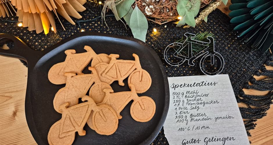 Black plate with bicycle-shaped cookies next to a handwritten recipe card and bike decoration. Festive wreath and paper fans in background.