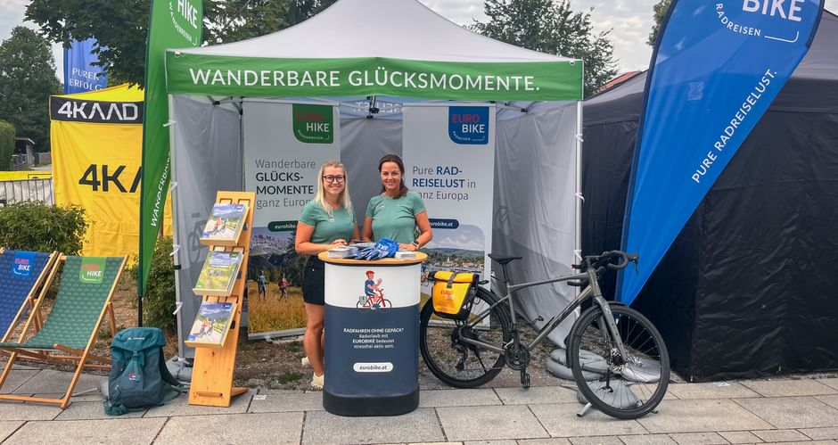 Two staff members in turquoise shirts at an Eurobike promotional tent with banners and a bicycle. Green banner reads 'Wanderbare Glücksmomente'.
