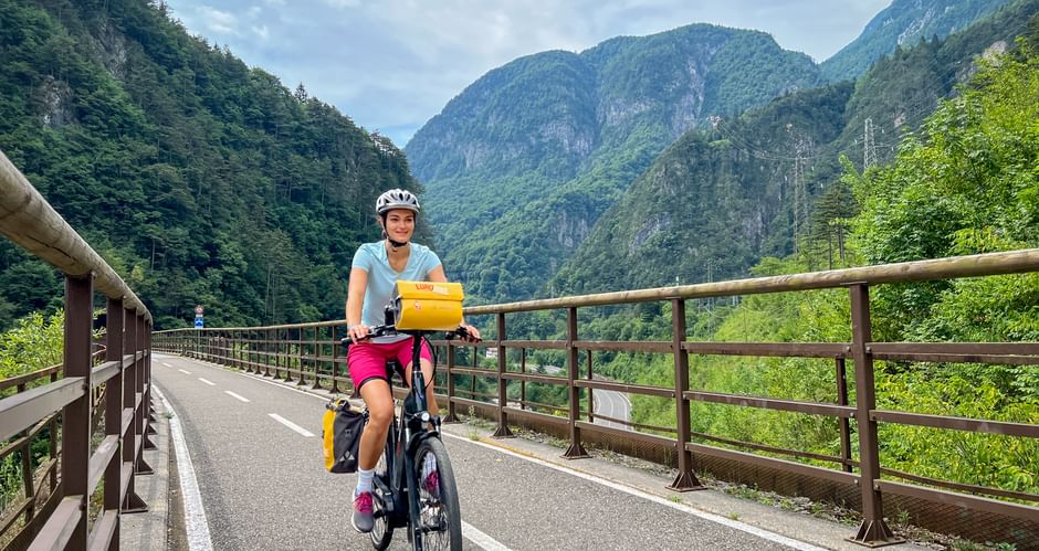 Radfahrerin mit gelben Packtaschen auf asphaltiertem Weg mit Metallgeländer, umgeben von bewaldeten Bergen im Alpental.