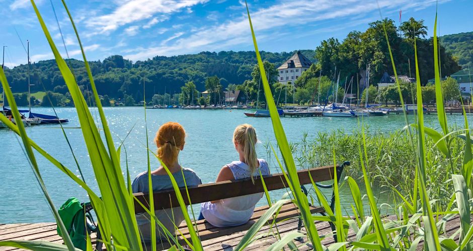 Two women sitting on a bench on a jetty in Weyer Bay with a view of Mattsee Castle