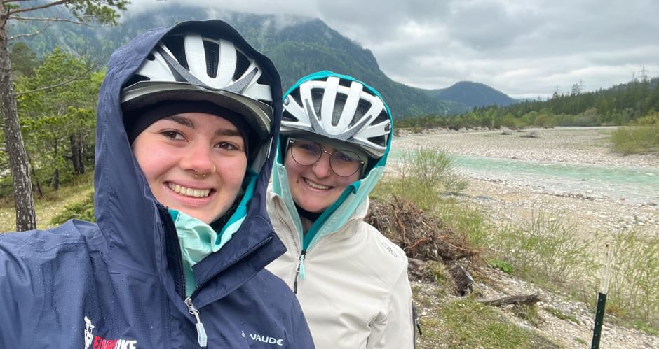 Two smiling cyclists in helmets and rain jackets taking a selfie by the Isar River with mountains and forest in the background.